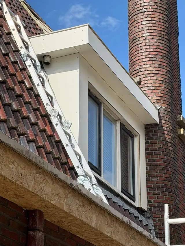 Nieuw wit afgewerkt houtwerk rond een raam in het pannendak, naast een ronde bakstenen schoorsteen