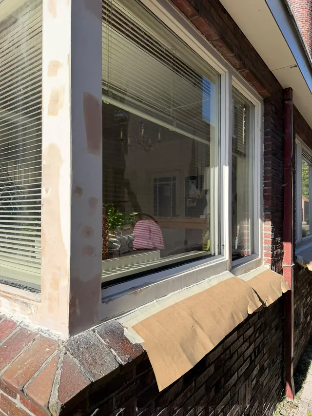 Hoek van een wit houten raamkozijn met lichte plamuurplekken na het herstel van houtrot, met afgeplakte vensterbank