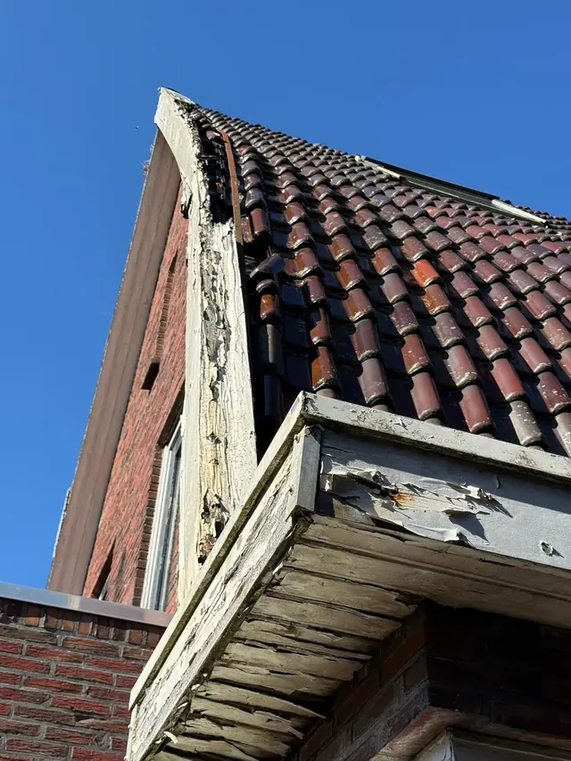 Verweerd houten boeiboord en overstek onder een rood pannendak, vóór het herstel